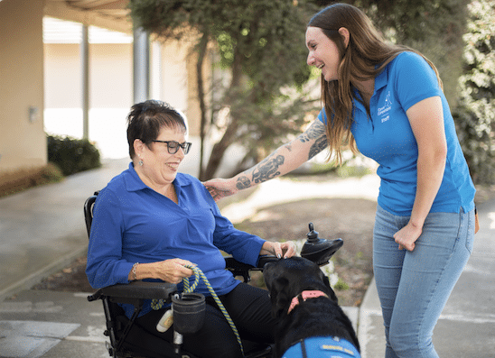 A woman in a wheelchair smiling and petting a service dog, while a standing woman in a blue polo shirt smiles and interacts with her.